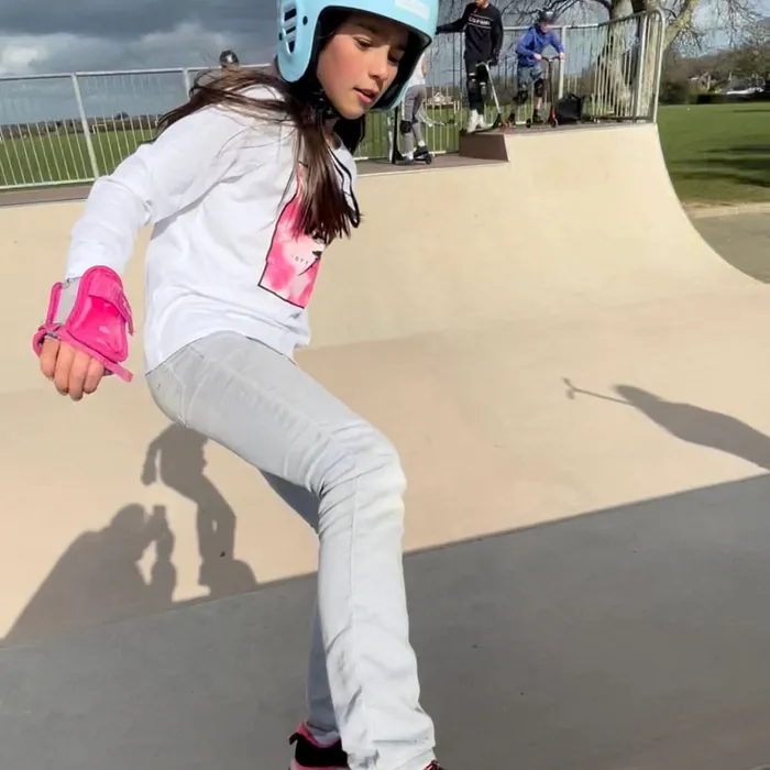 A young skater in a blue helmet and pink wrist guards doing a rock-fakey at a Dorset outdoor park