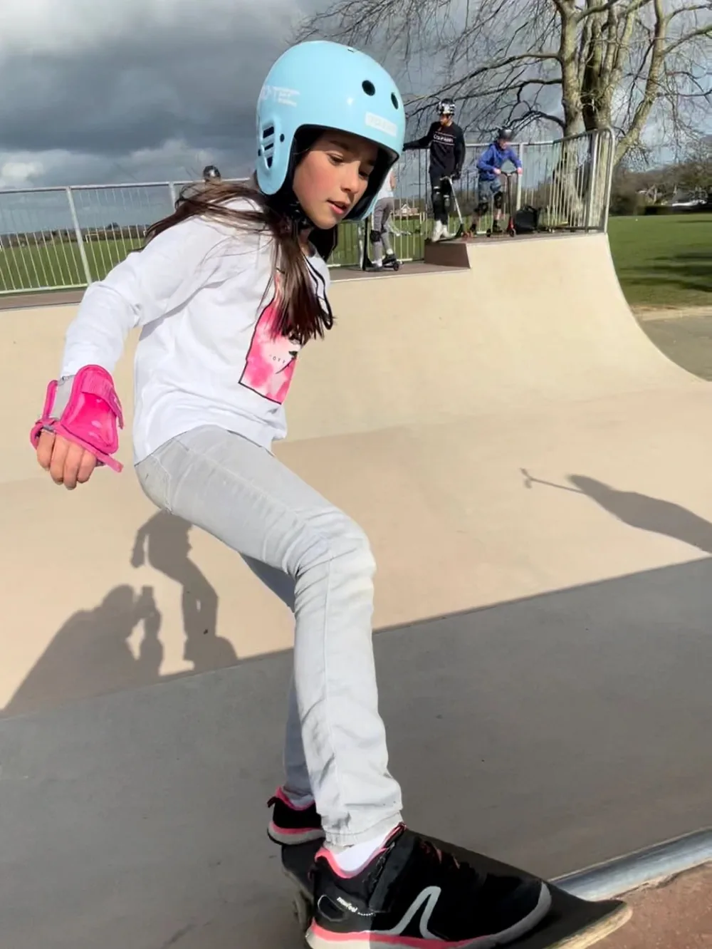 A young skater in a blue helmet and pink wrist guards doing a rock-fakey at a Dorset outdoor park